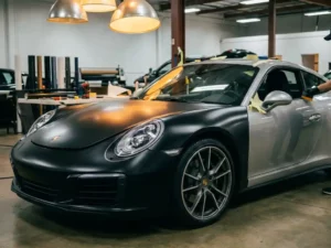 Sportwagen half ingepakt in matte zwarte vinyl folie naast originele metallic zilveren lak, in een industriële werkplaats met warm studiolicht.