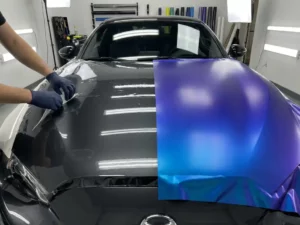 Handschoen van technicus vergelijkt kleurveranderende wrapfolie met transparante PPF-film op sportwagenmotorkap in professionele werkplaats.