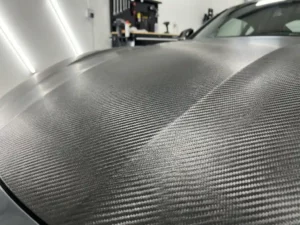 Sportswagenkap bekleed met carbon fiber vinyl, close-up van geweven textuur met metallic glans in professionele werkplaats.