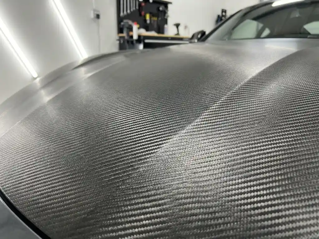 Sportswagenkap bekleed met carbon fiber vinyl, close-up van geweven textuur met metallic glans in professionele werkplaats.
