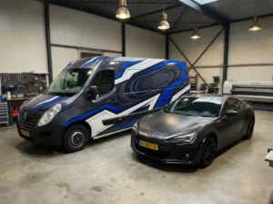 Beklede bezorgbus met levendige reclamefolie naast matte zwarte auto in modern Nederlands autobekledings atelier.