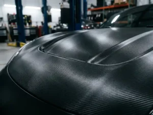 Sportswagen motorkap bekleed met zwart carbon fiber vinyl, geweven textuur zichtbaar in dramatisch studiolicht.