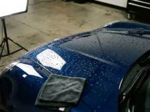 Keramisch gecoate motorklep van een sportwagen met waterparels op het glanzende donkerblauwe metallic oppervlak, microvezeldoek op de achtergrond.