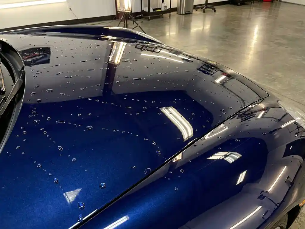 Glanzende motorkap met keramische coating waarop waterdruppels parelend afrollen, dramatische studiobelichting op diepblauw metallic oppervlak.