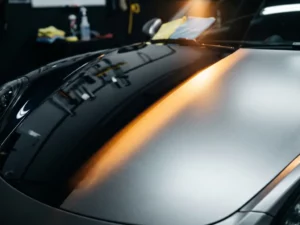 Sportwagenmotorkap half bedekt met matte PPF-folie, half met glanzende keramische coating, verlicht door amberkleurig studiolicht.