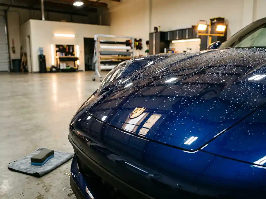 Glanzende motorkap van een sportwagen met keramische coating, waterdruppels op diepblauwe metallic lak, applicatiespons op microvezeldoek.