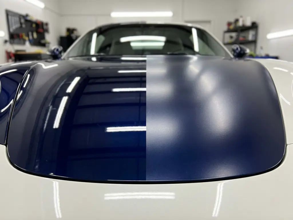 Sportwagenmotorkap half bedekt met keramische coating, half met matte PPF, onder professionele werkplaatsverlichting in marineblauw en parelwit.