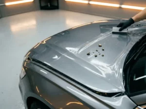 Transparante paint protection film aangebracht op zilveren motorkapglans, met grindstenen op het beschermde oppervlak in een werkplaats.