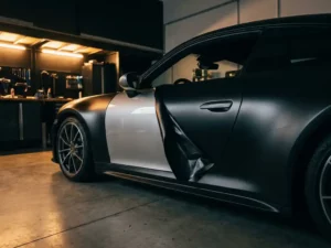Sportauto met matte zwarte vinylwrap, half afgestript met zichtbaar origineel zilver lak, gefotografeerd in moderne werkplaats.
